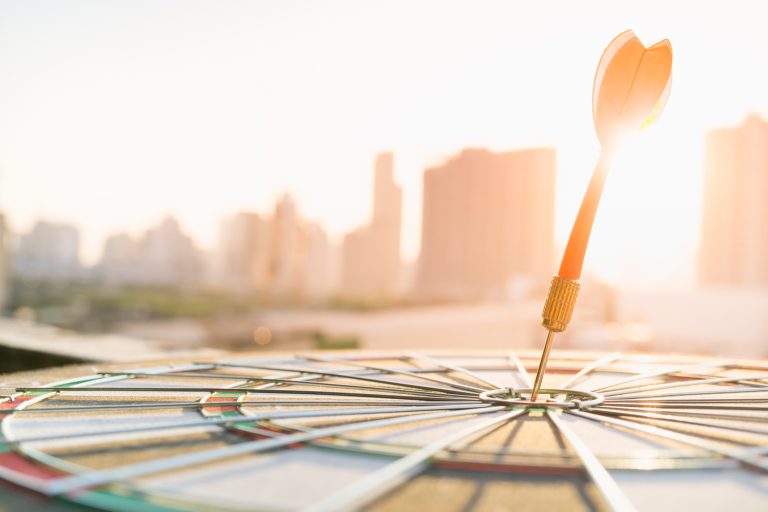 Red dart arrow hitting in the target center of dartboard with modern city and sunset background. Target business, achieve and victory concept .